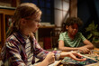 © pressmaster - Side view of serious youthful girl in eyeglasses looking at game board while sitting by table among her friends and throwing dice