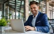 © OLGA - Joyful businessman in a blue suit working on a laptop in a modern office.
