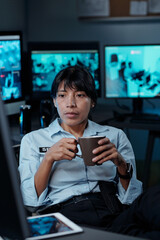 Wall Mural - Young serious woman with cup of coffee in hands looking at computer screen while sitting by workplace in surveillance room