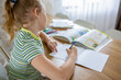 © evgeniia_1010 - A pretty school-age girl does her homework using a textbook and enters information into a workbook. The concept of combining online and home learning.