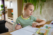 © evgeniia_1010 - A charming little girl does her homework based on a textbook and transfers the information to a workbook. The idea of combining online and home schooling.