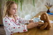 © evgeniia_1010 - A pretty blonde baby in cute pajamas with hearts is eating a delicious breakfast and feeding her beloved teddy bear. The concept of proper and balanced nutrition and games for children.