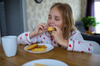 © evgeniia_1010 - A pretty blonde baby in cute pajamas is sitting at a comfortable table in the kitchen and enjoying a delicious and healthy breakfast. The idea of a proper and balanced diet.