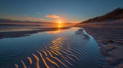The first rays of sunrise over a beach highlighting the ripples in the sand and creating a shimmering effect on the shallow water near the shore.