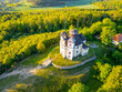 © pyty - An aerial view of the Church of Saint John the Baptist and Our Lady of Mount Carmel at Makova hora, Czechia, surrounded by lush greenery on a bright spring evening.