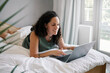 © Nicola Suttle/Stocksy - Smiling woman surfing internet via laptop on bed