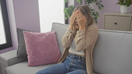 Poster - Young blonde woman sitting on sofa peeking in shock covering face and eyes with hand, looking through fingers with embarrassed expression. at home