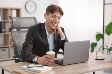 Wall Mural - Man with cup of coffee watching webinar at wooden table in office