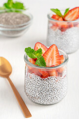 Homemade healthy chia pudding dessert made from seeds soaked in milk decorated with topping of fresh mint leaves and sliced ripe juicy strawberries served in glass bowl with spoon on white table