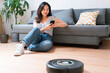 © nomad studio/Stocksy - Young woman using smartphone with robot vacuum cleaning floor