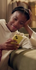 Poster - African woman, phone and texting by couch in home with smile, thinking or check notification in living room. Girl, smartphone and happy in night with typing by sofa with reading post on social media