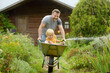 © Maria Sbytova - Happy little boy having fun in a wheelbarrow pushing by dad in domestic garden on warm sunny day. Child watering plants from a hose. Active outdoors games for kids in summer.