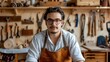 © Pnitha - Portrait of handsome young man wearing glasses and apron working in the workshop, sitting at workbench with tools on wall behind him. small business owner, local craftwork artist or carpenter professi