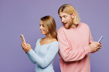 Wall Mural - Young couple two friends family man woman wearing pink blue casual clothes together hold use peek look at mobile cell phone stand back to back look camera isolated on pastel plain purple background.