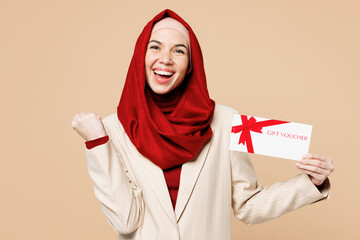 Wall Mural - Young Arabian Asian Muslim woman wear red abaya hijab suit clothes hold store gift coupon voucher card do winner gesture isolated on plain beige background. UAE middle eastern Islam religious concept.