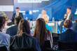 © Anton Gvozdikov - Business professionals in an audience attentively listening to a panel discussion at a conference with blurred speakers on stage.