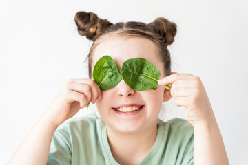 Wall Mural - Happy little girl of pre-teens, covering her eyes with spinach leaves. A girl in a green T-shirt on a white background is smiling with leaves in her hands. Concept: healthy eating