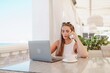 © svetograph - Woman coffee cafe laptop. Coffee break in cafe with sea view. Tranquil long haired woman drinking coffee in plant filled place. Woman sitting at a coffee shop with mobile phone drinking coffee.