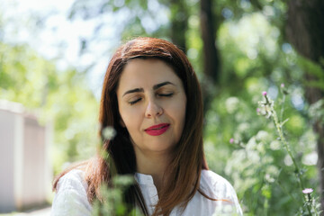 Wall Mural - Portrait of a woman about 45 years old with red lips, relaxed and happy with closed eyes with green background of foliage on a sunny day in the park. Concept of maturity, happiness and lifestyle