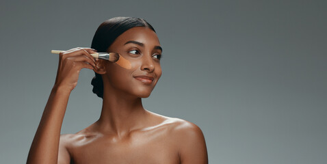 Wall Mural - Beauty, makeup and foundation brush of a woman with cosmetics, smile and dermatology. Portrait of a happy Indian model person with facial care, skin glow and wellness on a grey background in studio