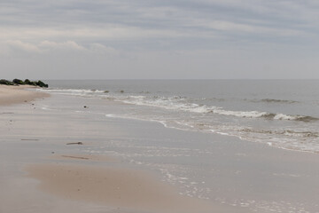 Wall Mural - This is an image of a beautiful day of the beach. The soft whitecaps of the waves coming in to batter the shore, carrying out the sand with each pass. The clouds are stretched across the sky.