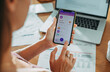© Marko Geber - Woman using banking app on smartphone with laptop and bills on desk