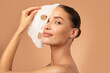 © Home-stock - Beautiful European lady removing moisturizing white fabric sheet mask from her face after using it, looking at camera, posing on beige background