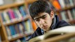 © mashimara - Back to school concept. Teenage boy with dark hair reading a book in a library, looking focused and serious, with shelves of books in the background.