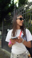 Wall Mural - A charming young brunette stands on the street, with a smartphone in her hands.