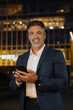 © Stock 4 You - Middle age entrepreneur businessman working on mobile cell phone. Smiling mature hispanic senior business man using app, holding smartphone cellphone at office building, looking at camera. Vertical