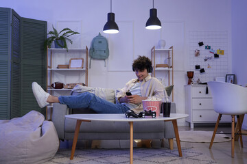 Canvas Print - Male student with headphones using mobile phone on sofa at home in evening