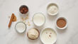 © Ron - Overhead view of ingredients for making milk, on a white background, with milk and cream in small bowls, cinnamon sticks, flax seeds, powdered sugar, cacao powder, vanilla powder