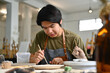 © saltdium - Two teenage Asian students focused on painting ceramic pieces in an art classroom together
