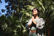 © insta_photos - Happy calm young Hispanic woman holding hands in namaste meditating doing yoga breathing exercises with eyes closed feeling peace of mind, mental balance standing in green nature tropical park.
