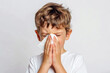 © Mirador - Young boy sneezing into a tissue with a plain white background. Represents common cold or allergy symptoms in a clean and simple setting.