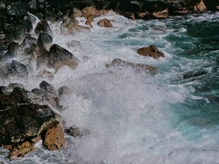 Wall Mural - Turquoise blue sea water with white foam on wave splashes. Water washes black volcanic rock stones. Tenerife, Puerto de la Cruz