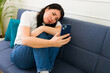 © AntonioDiaz - Worried young woman on a sofa, staring at her phone with concern, in a brightly lit living room setting