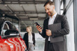 © anatoliycherkas - Smiling salesman using smartphone while having business call at car showroom. Car dealer holding clipboard in automobile showroom. Professional confident sales person working in modern car dealership.
