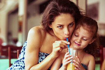 Wall Mural - Cute kid girl with happy beautiful mother drinking tasty healthy juice together with love in street summer restaurant. Closeup