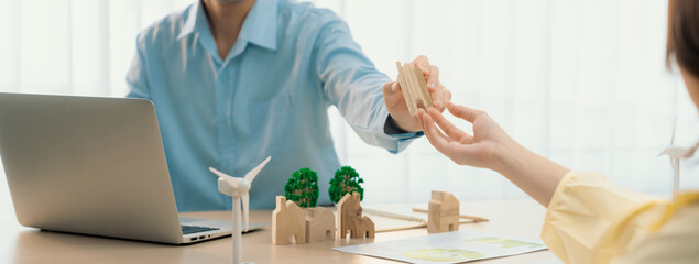 Wall Mural - Two business people talking about eco city at green business meeting on meeting table with windmill represented renewable energy, wooden house block and tree model represent green city. Delineation.