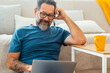 © simona - Adult man in casual clothing and eyeglasses using laptop and smiling while sitting on the floor against the sofa at home. Indoor technology computer leisure activity people lifestyle. Working online
