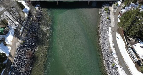 Wall Mural - Aerial of cars driving on river bridge surrounded by snowy forests, and mountain in the background