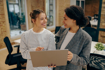 Two smiling female coworkers discussing project standing in office with laptop