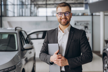 Wall Mural - Modern bearded man in glasses and suit vehicle sales consultant using laptop inside car dealership