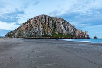 Wall Mural - Panorama of famous Morro Rock at dawn, Morro Bay, California. Sandy beach in the foreground; ocean to the right. Cloudy blue sky above.
