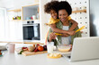 © Dorde - Playful young girl embracing her loving mother and holding spaghetti straps in both hands while making pasta for lunch, whipping ingredients in a bowl and using laptop for online cooking recipes.