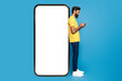 © Prostock-studio - A young Indian man wearing a yellow shirt and jeans is engrossed in using his smartphone. He is standing against a giant phone frame with a blank white screen, set against a vivid blue background