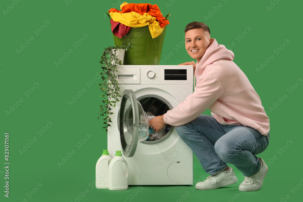 Handsome young man putting laundry into washing machine on green background
