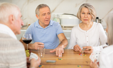 Wall Mural - Two senior married couples are having fun in cozy home atmosphere, playing card game poker. Player makes lead move and believes in luck, emotionally waiting for victory, calculate winning combination