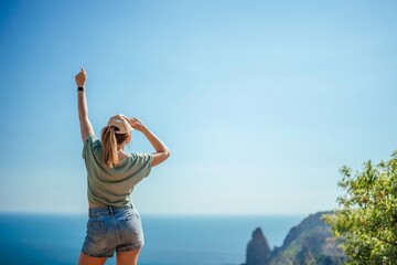 Wall Mural - Woman tourist sky sea. Happy traveler woman in hat enjoys vacation raised her hands up